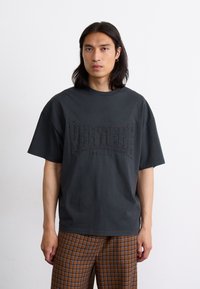 Dark gray cotton t-shirt with short sleeves, featuring textured logo "VERTIGO BERLIN" embroidered across the chest.