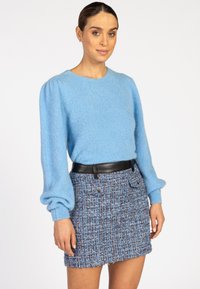 Vrouw in een lichtblauwe lange mouwen trui met een pofmouw en een blauwe tweed minirok met een zwarte riem, staande tegen een witte achtergrond.