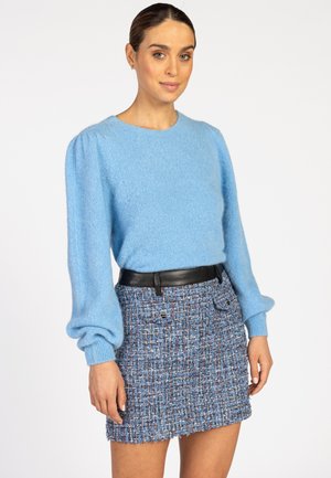 Frau in einem hellblauen langärmligen Pullover mit Puffärmeln und einem blauen Tweed-Minikleid mit schwarzem Gürtel, steht vor einem weißen Hintergrund.