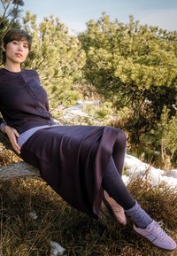 Knee-length, dark purple cardigan over a layered light top and dark leggings. Pink sneakers with textured gray socks, sitting on a log.
