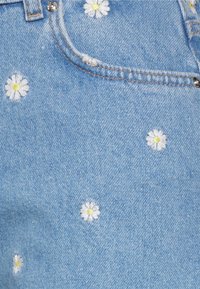 Ljusblå denimjeans med spridda vita och gula broderade tusenskönor. Har en framficka och silverfärgad hårdvara vid midjan.