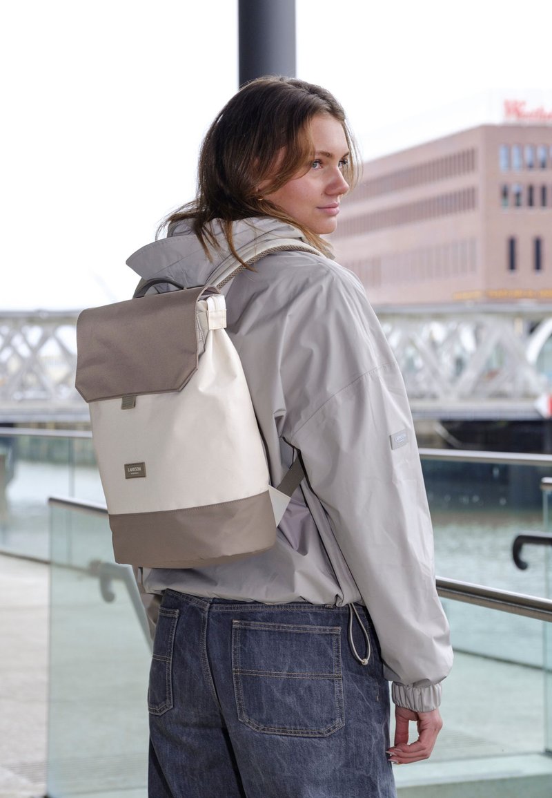 Jeune femme portant une veste grise et un jean, porte un sac à dos beige et gris à rabat roulant au bord de l'eau avec une rambarde en verre et un bâtiment en arrière-plan.