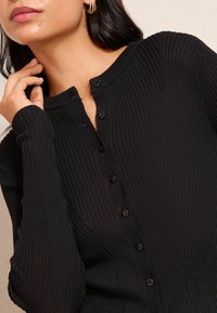 Schwarze gerippte Strickjacke mit Henley-Ausschnitt, ausgestattet mit sechs schwarzen Knöpfen und langen Ärmeln. Glatte Textur, taillierte Passform und dezente Details.