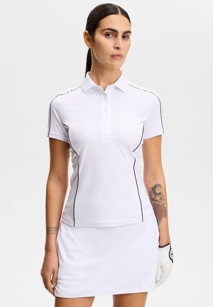 Donna che indossa una polo da golf bianca a maniche corte con bordatura nera, gonna da golf bianca e guanto da golf bianco, in piedi davanti a uno sfondo semplice.