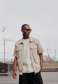 Jachetă bej cu mâneci scurte, cu două buzunare pe piept, purtată peste un tricou gri deschis; modelul poartă ochelari cu rame transparente și pantaloni negri.