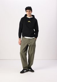 Black hoodie with front pocket and yellow logo, olive green wide-leg pants, black sneakers with white stripes, and black cap.