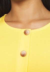 Žlutý oděv s kulatým výstřihem, třemi knoflíky a texturovaným povrchem. Knoflíky jsou světle hnědé s viditelným švem.