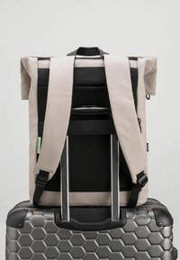 Rucsac gri cu capac rabatabil, cu spate din plasă neagră și curele ajustabile, plasat pe o valiză argintie cu model hexagonal. Design ergonomic vizibil.