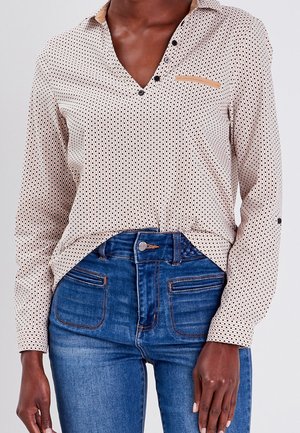 Donkergekleurd persoon met een beige blouse met patroon en knopen, licht ingestopt in een high-waisted blauwe jeans met steekzakken aan de voorkant.