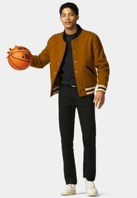 Bruine wolmix bomberjack met gestreepte manchetten, zwarte broek en witte sneakers. Houdt een oranje basketbal met het NBA-logo vast.