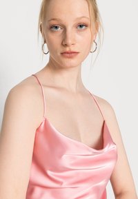 Camisola de cetim rosa com alças finas, apresentando um decote drapeado e uma textura suave e brilhante. Modelo apresenta um close-up.