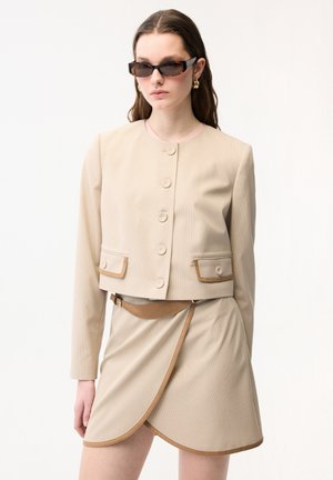 Vrouw draagt een beige cropped jack met knopen en een bijpassende wikkelrok met riem, gecombineerd met donkere zonnebril en gouden oorringen.