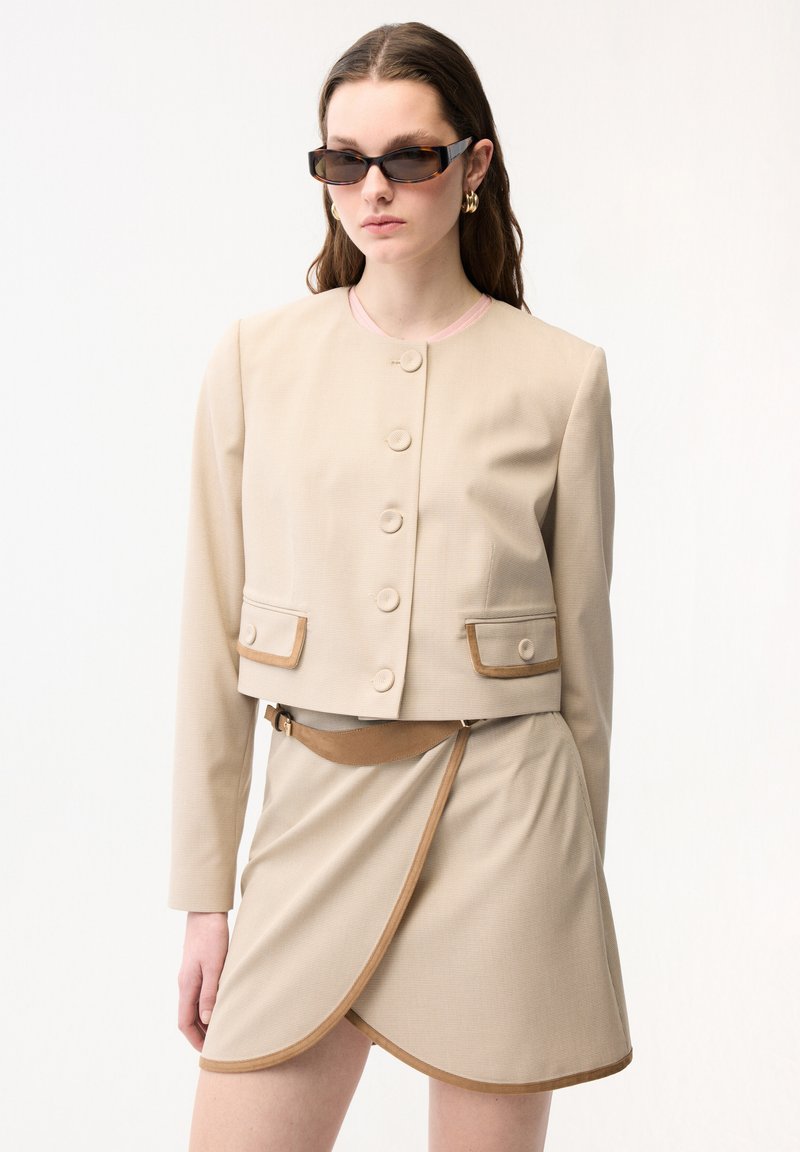 Donna che indossa una giacca beige corta con bottoni e una gonna a portafoglio coordinata con cintura, accessoriata con occhiali da sole scuri e orecchini a cerchio dorati.