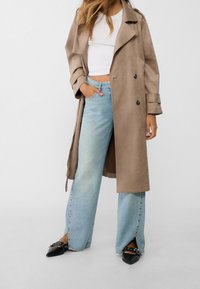 Beige suede trenchcoat met brede revers en een dubbelgeknopte ontwerpelement, gecombineerd met lichtblauwe wijde jeans met zijsplitten en zwarte puntige ballerina's.