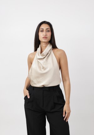 Vrouw met lang donker haar, die een beige mouwloos coltop en zwarte hoog getailleerde broek draagt, staande tegen een effen witte achtergrond.