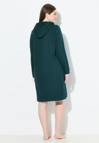 Vestido con capucha y mangas largas en verde oscuro. Fabricado con un tejido suave, de corte recto y con puños acanalados. Presenta un bolsillo tipo canguro en la parte frontal.