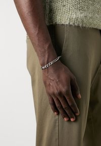 Pulsera de cadena de plata con eslabones gruesos y acentos puntiagudos, llevada en una muñeca desnuda, combinada con pantalones color verde oliva y un suéter texturizado.
