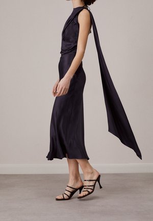 Kobieta w czarnej, satynowej sukience midi bez rękawów z długim, zwiewnym szalem z tyłu, w połączeniu z czarnymi, paskowymi sandałami na obcasie.