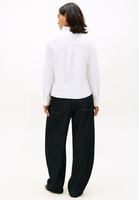 Žena stojící zády, oblečená v bílé blůze s dlouhými rukávy, volánovým límcem a širokými černými kalhotami.