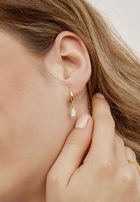 Nahaufnahme des Ohrs einer Frau, die einen goldenen Creolenohrring mit einem tropfenförmigen Perlenanhänger trägt und mit einer manikürten Hand ihr Haar berührt.