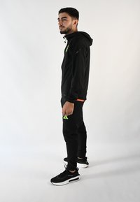 Chaqueta deportiva negra con logo verde, combinada con pantalones negros con rayas verdes. Zapatillas negras con suelas blancas.