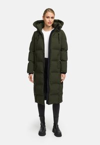 Lange, gewatteerde parka in donkergroen met een ritssluiting aan de voorkant, verstelbare capuchon en zijzakken, gedragen over een witte top en zwarte jeans. Zwarte laarzen.