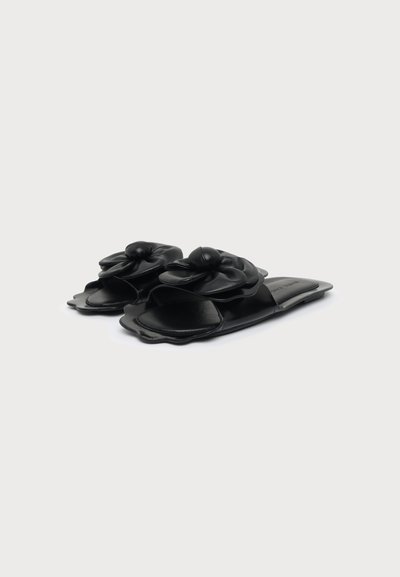 Sandales plates noires à bords festonnés avec une grande fleur décorative sur le dessus, présentées sur un fond blanc uni.