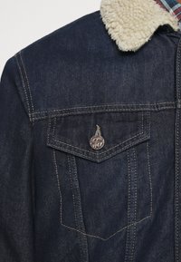 Giacca di jeans blu scuro con colletto in sherpa crema. Presenta dettagli con bottoni metallici e due tasche sul petto con patta a punta.