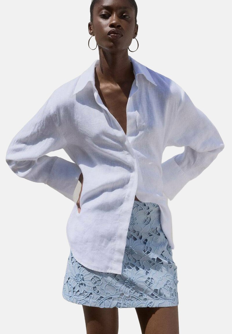 Camisa blanca de lino con mangas largas, con botones en la parte delantera y un corte relajado, combinada con una falda de encaje azul claro con patrones florales.