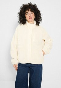 Kobieta z kręconymi włosami, ubrana w kremowy, puszysty żakiet i szerokie, ciemnoniebieskie dżinsy, pozująca z jedną ręką w kieszeni.