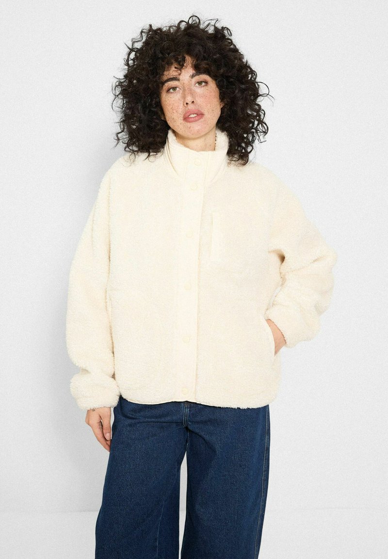 Kobieta z kręconymi włosami, ubrana w kremowy, puszysty żakiet i szerokie, ciemnoniebieskie dżinsy, pozująca z jedną ręką w kieszeni.