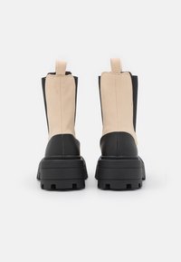 Botas de salto grosso com sola preta, parte superior em couro bege e laterais pretas. Apresentam uma alça para fácil calçar e uma sola robusta.