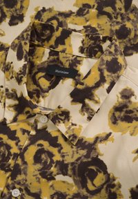 Chemise à manches courtes en tissu léger avec un motif abstrait jaune et marron, col chemise et fermeture par boutons blancs. Étiquette visible.