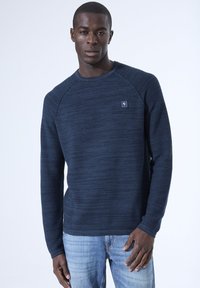 Marineblaues Sweatshirt mit langen Ärmeln, strukturiertem Design, geripptem Rundhalsausschnitt und kleinem quadratischen Logo auf der Brust, kombiniert mit hellen Jeans.