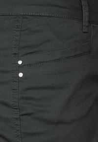Pantalones de mezclilla negros con una textura sutil, dos tachuelas decorativas plateadas y un bolsillo lateral con un diseño limpio y aerodinámico.