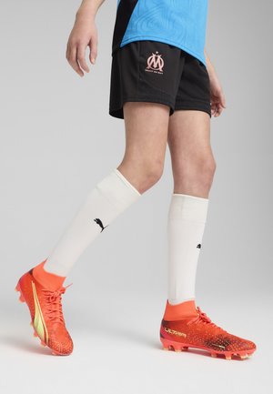 Erksad oranžid jalgpalliketsid, millel on gradientkujundus, koos mustade sportlike lühikeste pükstega, millel on roosa logo, ja valged sokid mustade detailidega.