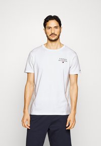 Hvit bomull t-skjorte med rund hals og korte ermer, med en svart logo trykket på venstre brystområde. Enkelt design.