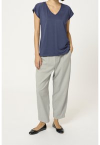 Donna che indossa un top blu a maniche corte con scollo a V, pantaloni larghi grigi chiari e ballerine nere in vernice, in piedi con una mano nella tasca.