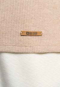 Tela beige texturizada con un patrón de gofre en la parte superior, sobre una tela suave de color crema. Se encuentra un etiquetado de cuero marrón con "BIG STAR" adjunto.