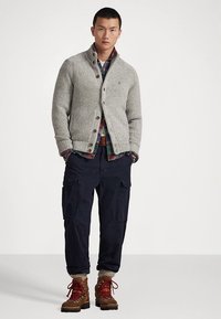 Gray knitted cardigan with a collar, brown buttons, layered over a plaid shirt. Navy cargo pants and brown boots complete the outfit.