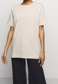 T-shirt in cotone beige con maniche corte, colletto rotondo, vestibilità rilassata e un piccolo logo vicino all'orlo. Abbinato a pantaloni larghi scuri.