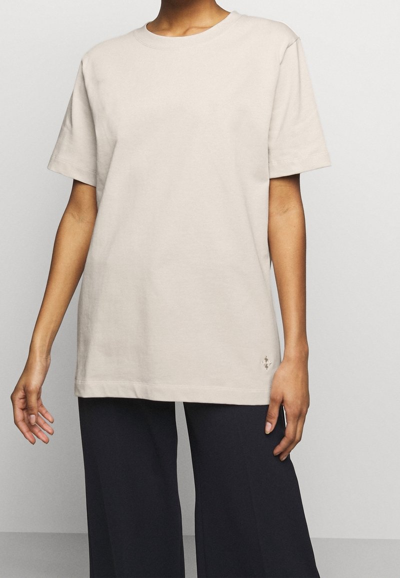 T-shirt in cotone beige con maniche corte, colletto rotondo, vestibilità rilassata e un piccolo logo vicino all'orlo. Abbinato a pantaloni larghi scuri.