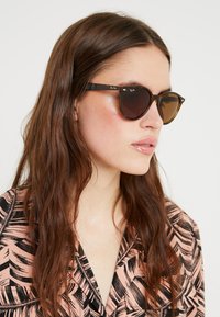 Ochelari de soare din țiglă cu lentile maro, accente negre și detalii metalice. Modelul poartă o cămașă roz cu un model.