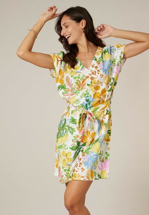 Femme souriante dansant dans une robe courte à ceinture, fleurie avec des motifs colorés jaune, vert, bleu et rose sur un fond clair.