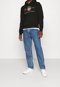Sudadera con capucha negra con un logo gráfico, combinada con vaqueros de mezclilla azul claros y zapatillas blancas. Ajuste casual con detalles en contraste.