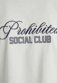 Tricou din bumbac alb cu inscripția "Interzis" în cursiv navy și "SOCIAL CLUB" în font bloc, ambele brodate pe față.
