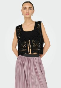 Gilet all'uncinetto nero con motivi floreali, design aperto sul davanti e chiusura a lacci. Indossato sopra una maglia nera e abbinato a una gonna plissettata lilla.