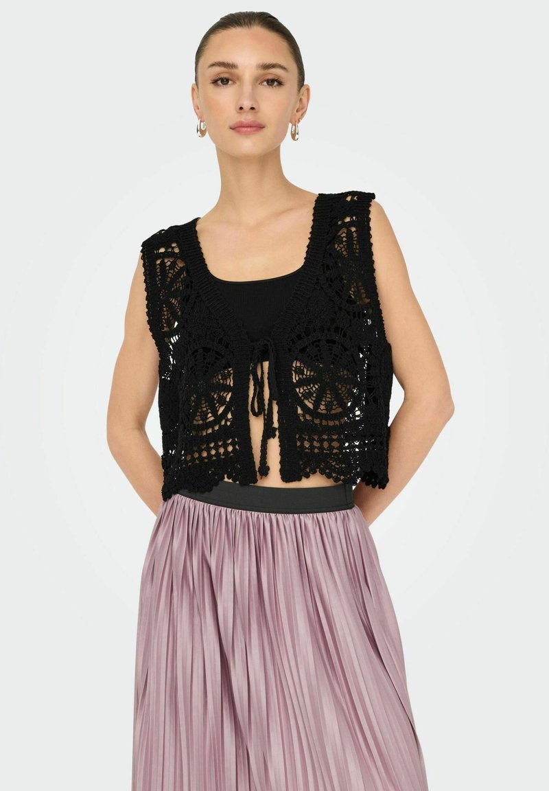 Gilet all'uncinetto nero con motivi floreali, design aperto sul davanti e chiusura a lacci. Indossato sopra una maglia nera e abbinato a una gonna plissettata lilla.