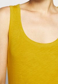 Lähikuva henkilöstä, jolla on päällä olkahihnaton sinapinkeltainen toppi, joka paljastaa kaulan, hartian ja ylärinnan.