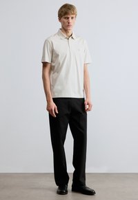 Béžová bavlnená polokošeľa s krátkym rukávom, golierom a malým logom na ľavej strane. Nosí sa s čiernymi nohavicami a čiernymi topánkami.